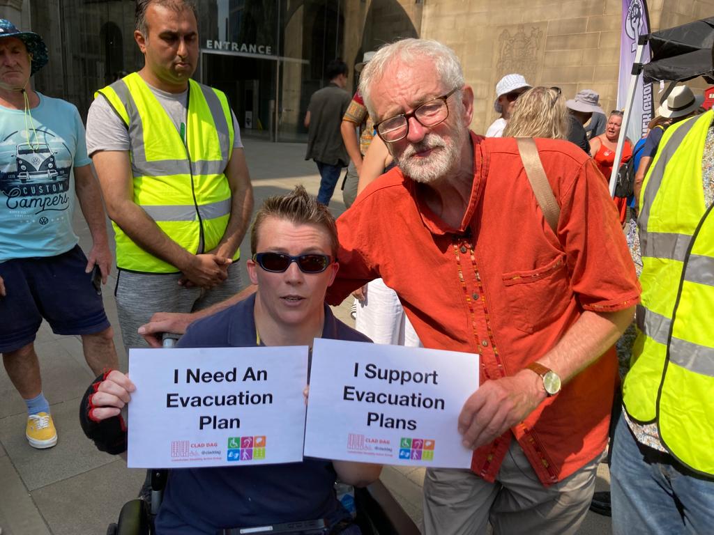 Jeremy is stood next to a wheelchair user in a sunny public square holding a "I support evacuation plans" sign