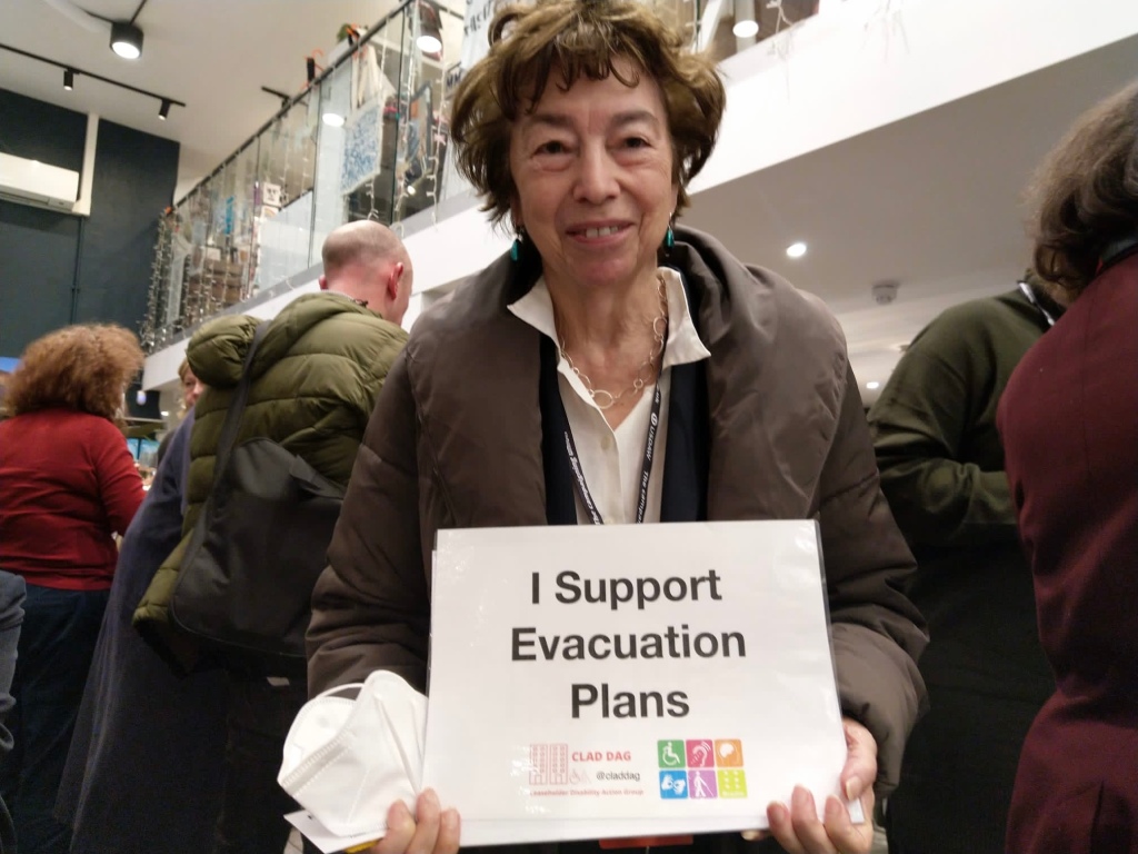 Nicky is in a public space smiling at the camera in a coat holding a sign which reads "I support evacuation plans"
