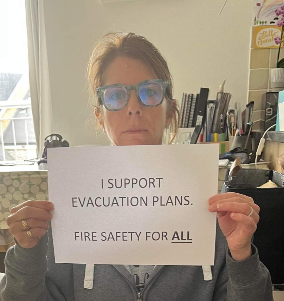 Liz is sitting in the kitchen looking serious and holding a sign which reads "I support evacuation plans"