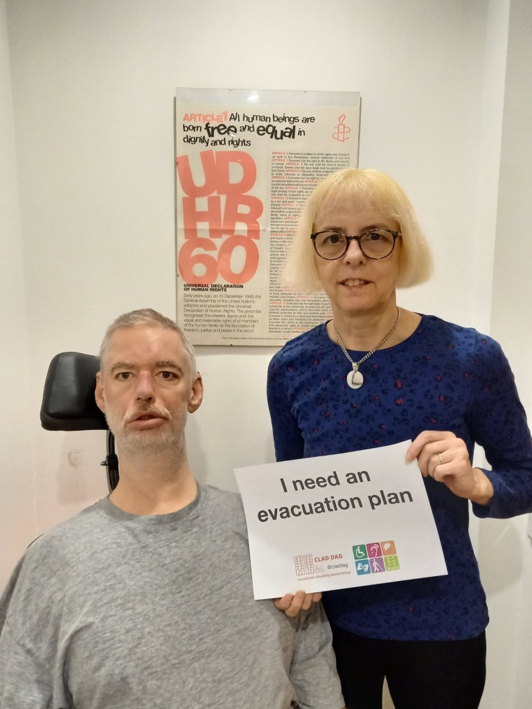 Alan is seated in his wheelchair, next to a woman stood next to him holding a sign which reads "I need an evacuation plan"