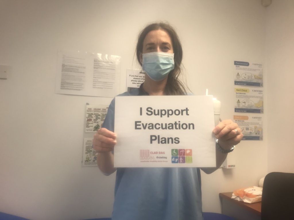 A female doctor in a face covering is stood in a GP surgery holding up a sign which reads "I support evacuation plans"