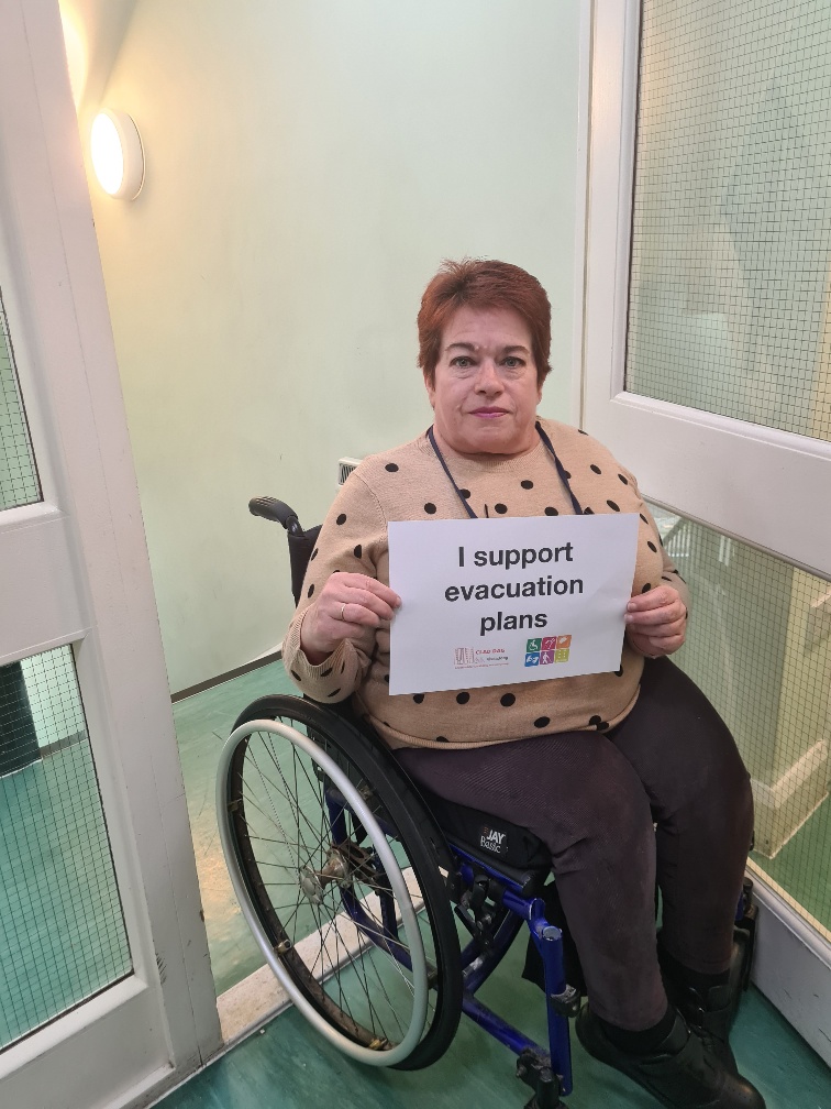 Rosemary, a wheelchair user, is a stairwell of a residential building holding a sign which reads "I support evacuation plans"